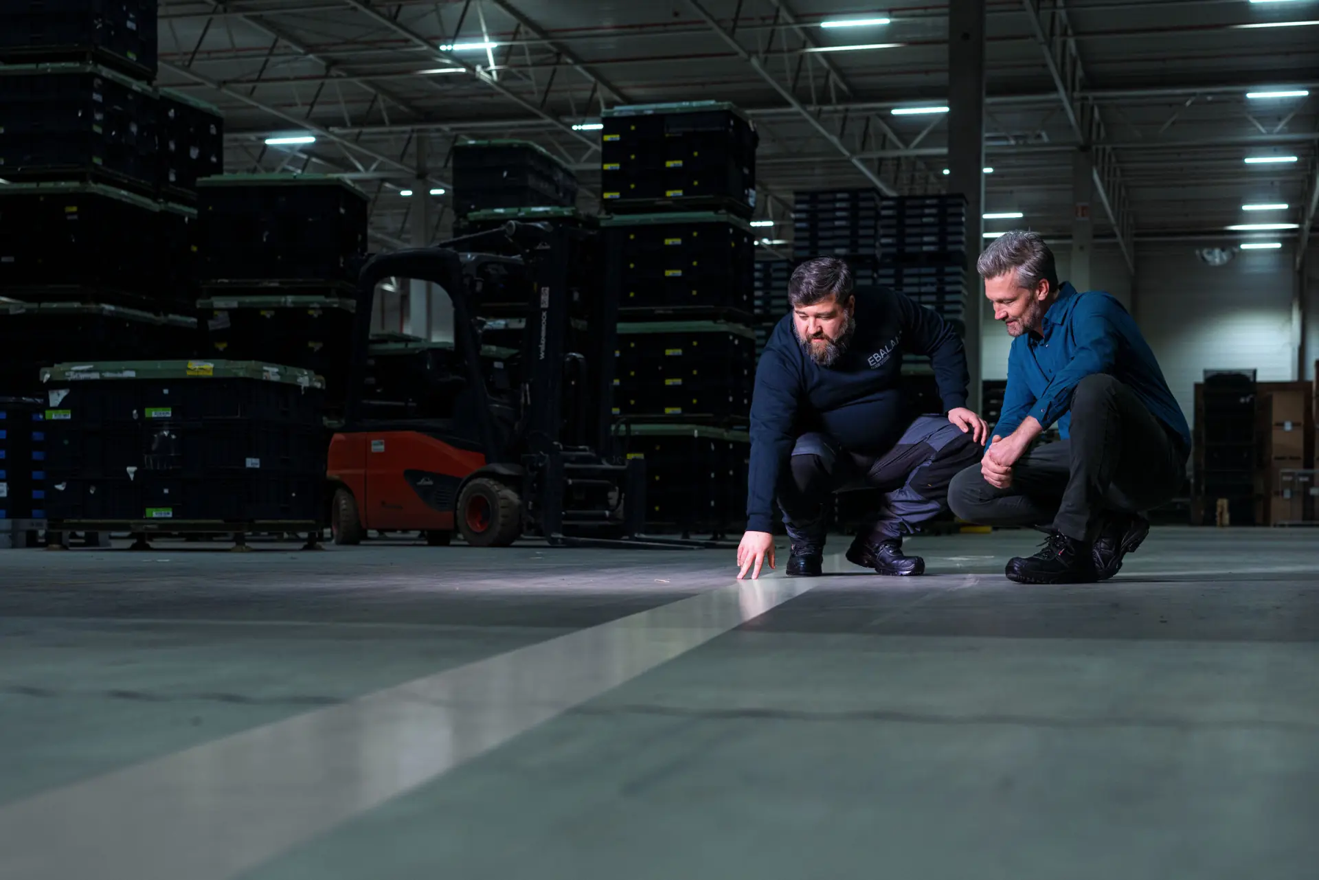 Zwei Männer prüfen eine sanierte EBALAN POWERFLEX Fuge im Fahrweg einer Logistikhalle bei DLG in Duisburg, im Hintergrund ein Stapler